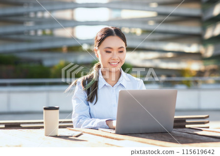 A young Asian woman is sitting at a table, focused on her laptop screen. Her hands are typing on the keyboard as she works or studies. The room appears well-lit, with minimalistic decor. A young Asian woman is sitting at a table, focused on her laptop screen. Her hands are typing on the keyboard as she works or studies. The room appears well-lit, with minimalistic decor. 115619642