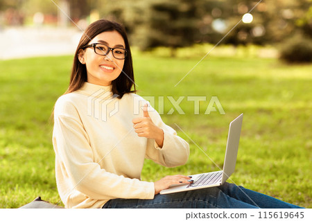 Asian lady is seen sitting on the grass in a sunny park during the afternoon. She is wearing a cream sweater and glasses, working on her laptop while smiling and giving a thumbs-up. 115619645
