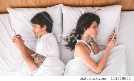 A man and a woman are lying in bed, each using their smartphones. The couple is lying on their sides, facing away from each other. The image is taken from above, looking down on the couple. 115619662