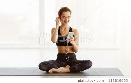 A woman is seated on a yoga mat, listening to music with headphones on. She appears focused and relaxed as she immerses herself in the music. A woman is seated on a yoga mat, listening to music with headphones on. She appears focused and relaxed as she immerses herself in the music. 115619663
