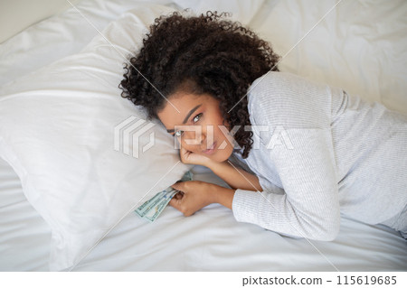 A young Hispanic woman with curly hair is lying on her stomach on a soft, white bed, her head propped on her hand while she smiles at the camera and playfully holds a few dollar bills 115619685