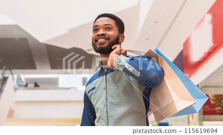 Positive black man is seen holding multiple shopping bags while walking through a bustling mall. He appears focused on his purchases, maneuvering through the crowd with ease. 115619694