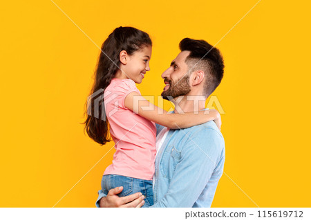 Father's love. Loving father holding his cute little daughter, dad bonding with her child girl, having fun with kid over yellow background, side view, copy space Father's love. Loving father holding his cute little daughter, dad bonding with her child girl, having fun with kid over yellow background, side view, copy space 115619712