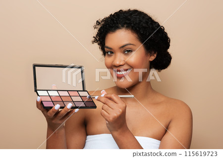 A young woman is shown holding a colorful makeup palette in her hand, examining the different shades and products. She appears focused on selecting the right colors for her look. 115619723