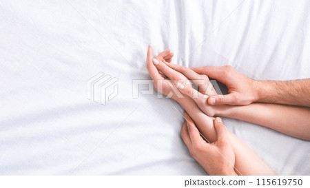 This image shows the hands of a couple intertwined on a white bed, suggesting intimacy and connection, cropped, copy space 115619750