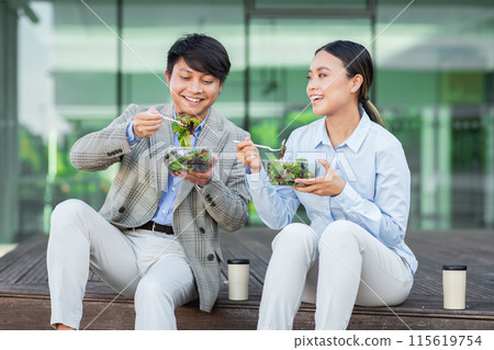 Two Asian individuals, dressed in business attire, are seen seated at a table. Both are actively engaged in consuming a bowl of salad, showcasing a healthy meal choice during their work break. 115619754