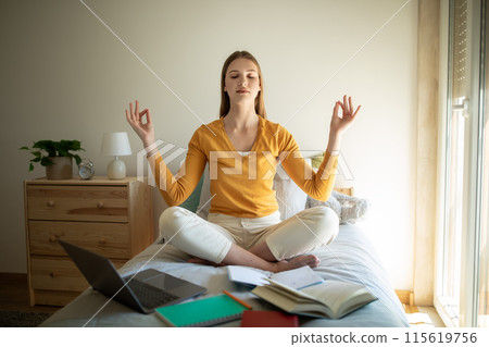 A teen girl is sitting on her bed, meditating with closed eyes and a serene expression. She is surrounded by open books, a laptop, and notebooks, indicating she is taking a break from studying 115619756
