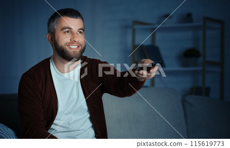 A bearded man sits on a couch, smiling while pointing a remote control towards a television in a cozy living room at night. 115619773