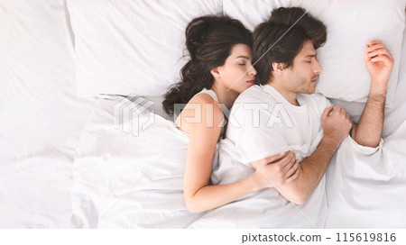 A couple is sleeping peacefully in bed together, with the woman nestled against the mans back, wearing white shirts and are completely relaxed, suggesting a comfortable and loving relationship. 115619816