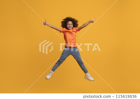 African American girl jumping in the air with her arms outstretched and giving a thumbs up with both hands. She is smiling and appears happy. The background is a solid yellow color. 115619836