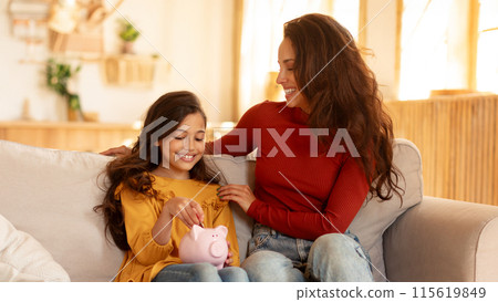 Kid And Money, Personal Savings. Happy Arab Mother And Preteen Daughter Sitting With Piggybank, Girl Putting Coin, Caring For Finances Safety And Future Abundance, Sitting On Couch At Home 115619849