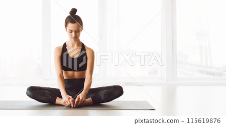 A serene young woman is sitting on a yoga mat in the lotus position with her eyes closed. She is in a well-lit studio with large windows, and the soft morning light accentuates the calm atmosphere 115619876