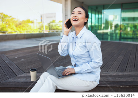 Asian woman is seated on a bench, engaged in conversation on her phone, holding computer on her lap. She appears focused, possibly discussing important matters 115620014