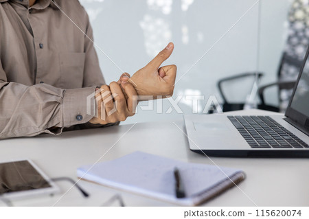 Businessman holding his wrist from pain while working on a laptop at the office. Workplace injury, office ergonomics, and healthcare. Businessman holding his wrist from pain while working on a laptop at the office. Workplace injury, office ergonomics, and healthcare. 115620074
