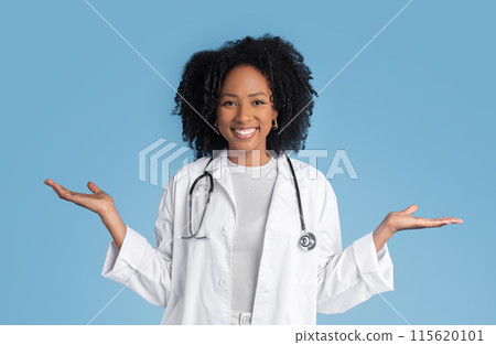 Cheerful young black lady doctor therapist in white coat hold free space in hands, isolated on blue studio background. Ad and offer, professional praxis, choice of health care 115620101