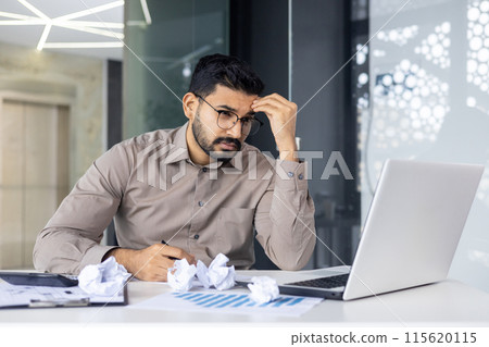 Frustrated businessman sitting at desk with crumpled papers, analyzing documents on laptop in modern office environment. 115620115