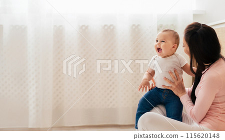 A woman is seated, cradling a baby in her lap. She gazes down lovingly at the infant, who appears calm and content in her arms, panorama with copy space A woman is seated, cradling a baby in her lap. She gazes down lovingly at the infant, who appears calm and content in her arms, panorama with copy space 115620148