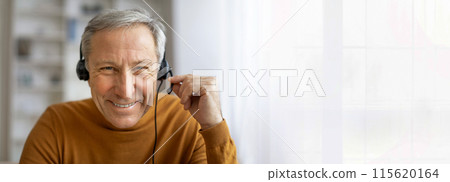 A senior man with gray hair is wearing a headset and smiling at the camera. He is likely working from home, as he is sitting in front of a window, copy space A senior man with gray hair is wearing a headset and smiling at the camera. He is likely working from home, as he is sitting in front of a window, copy space 115620164
