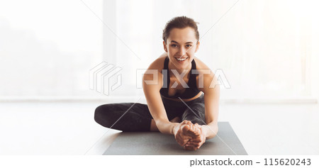 A woman is sitting on a yoga mat, looking directly at the camera with a smile on her face. She appears relaxed and content in her yoga practice. 115620243