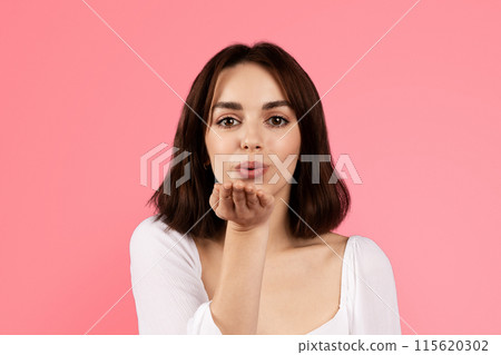 Happy millennial caucasian brunette woman making blowing kiss with hand, isolated on pink background, studio. Flirt, beauty care, holiday, lifestyle, ad and offer, love and romance 115620302