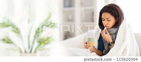 African American woman wrapped in a blanket sits on a couch, holding a mug of liquid in one hand while taking a pill with the other. She looks tired and unwell, copy space 115620304
