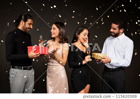 Two multiracial couples beautiful young men and women in beautiful outfits celebrating Christmas together on black background, exchanging xmas gifts, drinking champagne. New Year celebration 115620309
