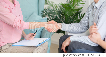 A therapist and a client shake hands during a consultation in a comfortable, warmly-lit office. The therapist holds a clipboard and pen, cropped, panorama 115620318
