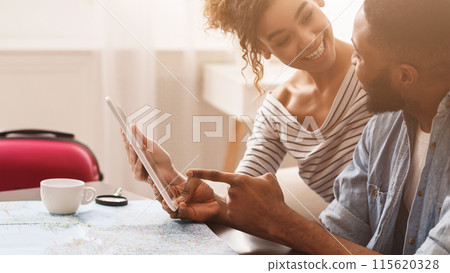 Black man and woman are seated at a table, engaged with a tablet device. The man and woman are focused on the screen, appearing to discuss or work together on the digital device. 115620328