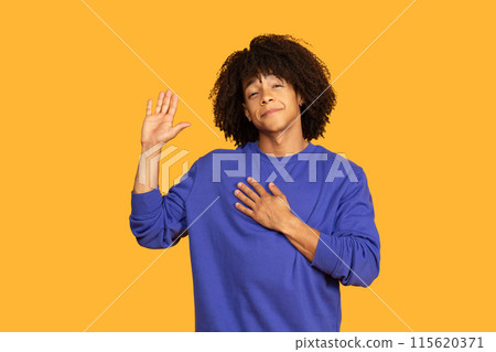A young man with curly black hair, wearing a blue sweatshirt, is standing against a yellow background. He is waving with his right hand and placing his left hand over his chest, smiling gently. A young man with curly black hair, wearing a blue sweatshirt, is standing against a yellow background. He is waving with his right hand and placing his left hand over his chest, smiling gently. 115620371