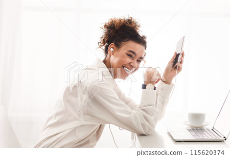 African American cheerful young woman with headphones is tilting her head back in laughter, holding a smartphone, and clearly enjoying a moment of music or conversation African American cheerful young woman with headphones is tilting her head back in laughter, holding a smartphone, and clearly enjoying a moment of music or conversation 115620374