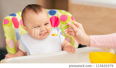 A woman is sitting and feeding a baby with a spoon. The woman hand is holding the spoon, while the baby is looking at the spoon. The baby seems to be enjoying the feeding process. 115620379