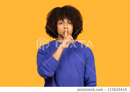 A man with curly black hair is wearing a blue sweatshirt and gesturing for silence with his finger to his lips against a yellow background. A man with curly black hair is wearing a blue sweatshirt and gesturing for silence with his finger to his lips against a yellow background. 115620416