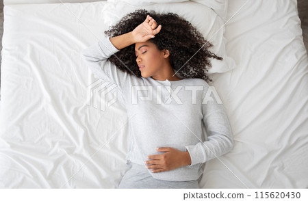 A young Hispanic woman in a tranquil state of sleep, lying comfortably in a bed adorned with white linens. Her pose suggests a deep and restful slumber, with one arm resting gently on her forehead 115620430