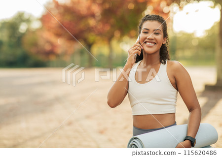 A cheerful woman dressed in activewear is seen smiling and talking on a smartphone while holding a rolled-up yoga mat. She is standing outdoors in a park with autumn foliage A cheerful woman dressed in activewear is seen smiling and talking on a smartphone while holding a rolled-up yoga mat. She is standing outdoors in a park with autumn foliage 115620486