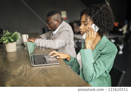 Business Communication. African American Businesswoman Talking On Phone Typing And Surfing Web On Laptop Sitting At Workplace Near Colleague Man In Modern Office, Side View, Selective Focus 115620551