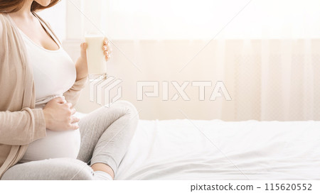 A pregnant woman is sitting on a white bed and enjoying a glass of milk while gently touching her belly. Soft natural sunlight filters through sheer curtains, creating a calming atmosphere, cropped 115620552