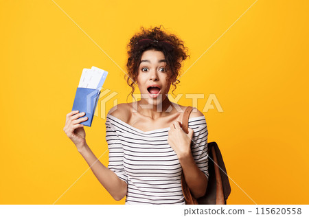 A joyful young African American woman with curly hair stands against a vivid yellow backdrop, holding what appears to be a pair of airline tickets and a passport A joyful young African American woman with curly hair stands against a vivid yellow backdrop, holding what appears to be a pair of airline tickets and a passport 115620558
