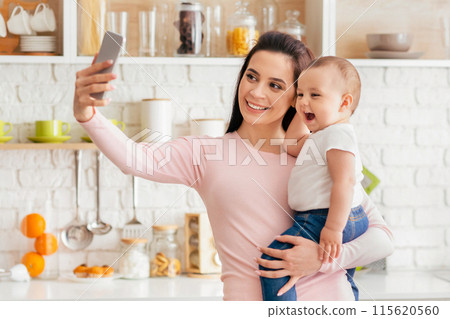 A woman holding her baby in her arms while taking a selfie with a smartphone. The mother and child are smiling at the camera in a casual and joyful moment. 115620560