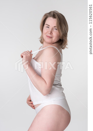 Serious woman in white underwear puts hand on stomach while holding strap of camisole. Overweight middle aged adult woman looking over shoulder at camera, represents women's healthy lifestyle concept Serious woman in white underwear puts hand on stomach while holding strap of camisole. Overweight middle aged adult woman looking over shoulder at camera, represents women's healthy lifestyle concept 115620561