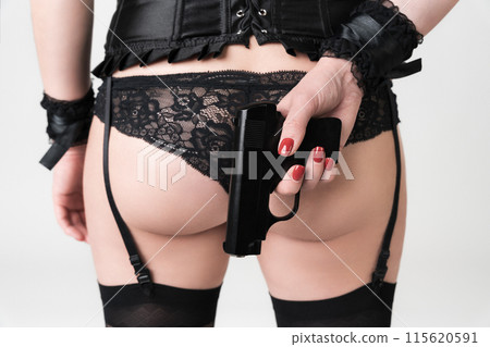Rear view of femme fatale holding gun in hand, selective focus on pistol, with close-up of buttocks in black lace panties and stockings. Unrecognizable real female body in lingerie on white background 115620591