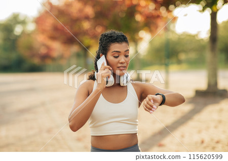 A young woman dressed in athletic clothing is in a park during the autumn season, talking on smartphone, with wireless headphones around her neck, while simultaneously glancing at her smartwatch. 115620599