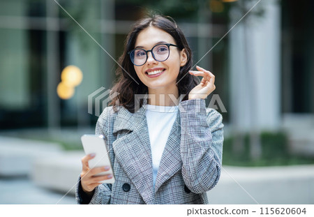 Business App. Happy Young Businesswoman Using Mobile Phone Wearing Glasses And Smiling At Camera, Scrolling Social Media Feed And Reading Business News Online Standing In Urban Area Outdoor 115620604