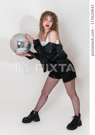 Confident woman holding disco mirror ball and looking at camera, standing on white. Beautiful young adult pin up girl with long curly hair dressed in black jacket, shorts and boots in clubbing style Confident woman holding disco mirror ball and looking at camera, standing on white. Beautiful young adult pin up girl with long curly hair dressed in black jacket, shorts and boots in clubbing style 115620642