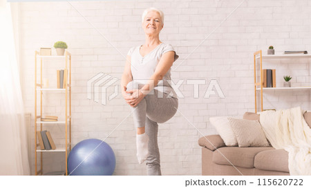 In a living room, senior woman is seen practicing a yoga pose. She is focusing on her balance and flexibility as she stretches her body 115620722