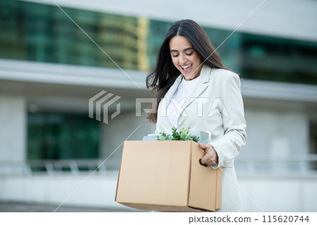 A young businesswoman, dressed in a white blazer and white shirt, walks away from her office building while carrying a cardboard box filled with her belongings A young businesswoman, dressed in a white blazer and white shirt, walks away from her office building while carrying a cardboard box filled with her belongings 115620744