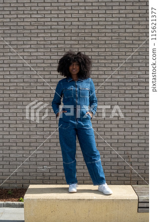 Confident woman in denim jumpsuit, against brick wall, showcasing stylish fashion sense Confident woman in denim jumpsuit, against brick wall, showcasing stylish fashion sense 115621377