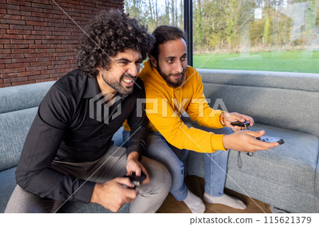 Two friends are energetically playing video games together on a comfortable couch in a living room 115621379