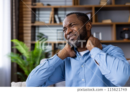 A man is sitting indoors, visibly suffering from neck pain. He is holding his neck with a pained expression, indicating discomfort. 115621749