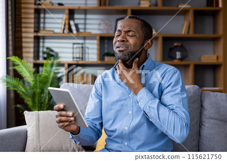 Man experiencing throat pain while consulting a doctor online via video call in a home office setting. Holding a tablet and displaying discomfort. 115621750