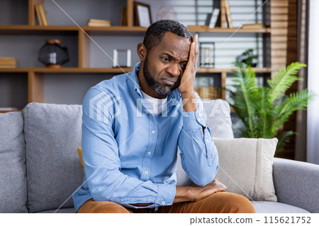 Man sitting on a couch looking stressed and worried, conveying feelings of anxiety and concern in a modern living room setting. 115621752